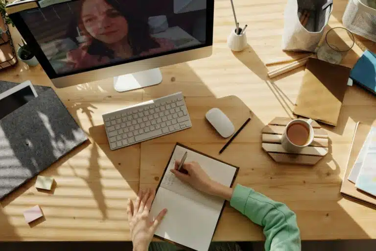 Aerial shot of people taking notes while watching a computer screen on a desk, stationery and a coffee cup are on the desk