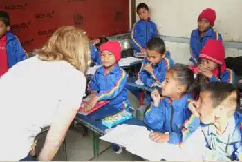 An older teacher teaching English to young learners in Nepal. The students are seated at their desks.